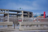 An urban landscape featuring multiple overpasses and highway bridges. The foreground shows a concrete barrier with metal railings, while towering over the scene are elevated roadways. In the background, there are city buildings, notably including a prominent red cylinder-shaped structure next to modern glass and steel buildings.