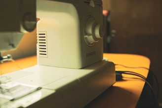 A close-up view of a sewing machine positioned on a yellow surface. The machine is white and appears to be a Singer brand. Various details of the machine, such as the dials and stitching area, are clearly visible. The background is slightly blurred and out of focus.