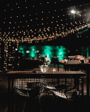A warmly lit outdoor setting features a table with wire chairs and a vase of flowers. String lights create a bokeh effect above, while a soft green illumination bathes the background.