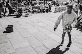 A street performer is captured in mid-movement, wearing a sweater, plaid shorts, and roller skates, with a decorative necklace. He performs on a paved surface, while an audience watches from benches and chairs. A cardboard box is on the ground nearby, possibly for collecting tips.