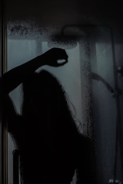 A silhouette of a person is visible behind a fogged-up glass shower door, with some streaks and droplets on the glass. The scene is dimly lit, adding a mysterious or introspective atmosphere.