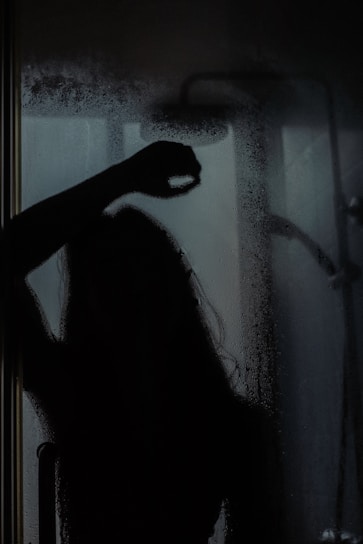 A silhouette of a person is visible behind a fogged-up glass shower door, with some streaks and droplets on the glass. The scene is dimly lit, adding a mysterious or introspective atmosphere.