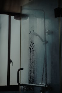 A shower glass door with water droplets and a distinct handprint in the condensation. A showerhead and window are visible in the background, giving the setting a gloomy or mysterious ambiance.