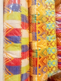 Colorful woven fabric rolls featuring intricate patterns and bright geometric designs. The fabrics display a mix of red, yellow, blue, and orange hues with various shapes such as diamonds and lines, creating a vibrant appearance.