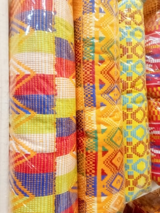 Colorful woven fabric rolls featuring intricate patterns and bright geometric designs. The fabrics display a mix of red, yellow, blue, and orange hues with various shapes such as diamonds and lines, creating a vibrant appearance.
