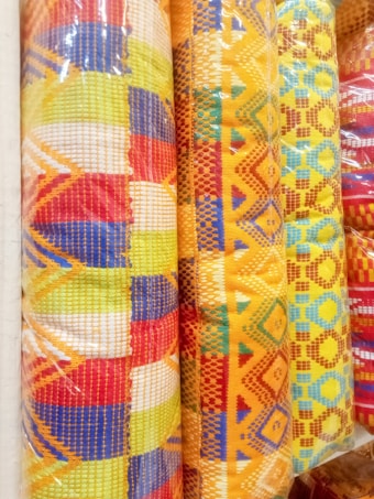 Colorful woven fabric rolls featuring intricate patterns and bright geometric designs. The fabrics display a mix of red, yellow, blue, and orange hues with various shapes such as diamonds and lines, creating a vibrant appearance.