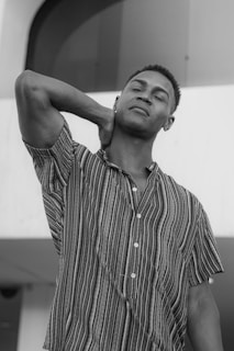 A person stands with a relaxed expression, one arm raised to the neck. Wearing a short-sleeved, button-down shirt with vertical stripes, the individual appears to be posing in front of a large, curved structure.