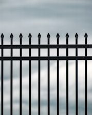 A vertical iron fence with pointed tops is prominently displayed against a cloudy sky backdrop. The bars are evenly spaced and have a sleek, black finish that contrasts with the pale, overcast sky.