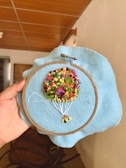A hand is holding an embroidery hoop with a piece of blue fabric stretched in it. The embroidery depicts a colorful hot air balloon with a bunch of multicolored knots representing the balloon and white threads representing the ropes attaching it to the basket. The background is an indoor setting with wooden furniture and a tiled ceiling visible.