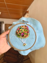 A hand is holding an embroidery hoop with a piece of blue fabric stretched in it. The embroidery depicts a colorful hot air balloon with a bunch of multicolored knots representing the balloon and white threads representing the ropes attaching it to the basket. The background is an indoor setting with wooden furniture and a tiled ceiling visible.