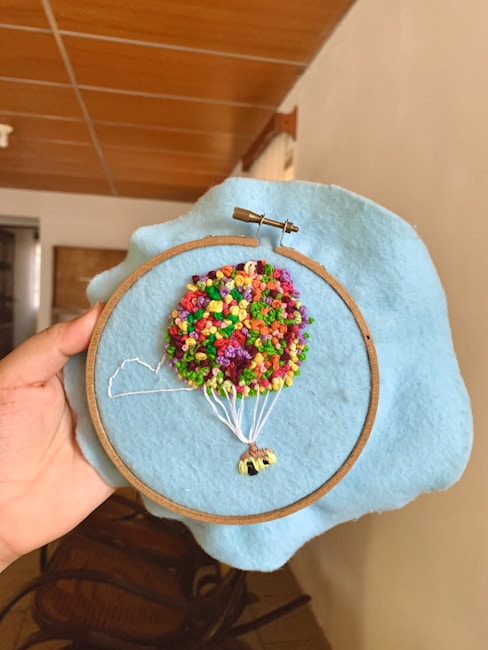 A hand is holding an embroidery hoop with a piece of blue fabric stretched in it. The embroidery depicts a colorful hot air balloon with a bunch of multicolored knots representing the balloon and white threads representing the ropes attaching it to the basket. The background is an indoor setting with wooden furniture and a tiled ceiling visible.