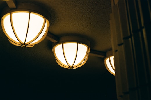 Three spherical ceiling lights with a warm glow illuminate a textured ceiling. The lights are encased in a decorative metal grid, suggesting an industrial or vintage style. The setting appears dim, enhancing the focus on the glowing lights.