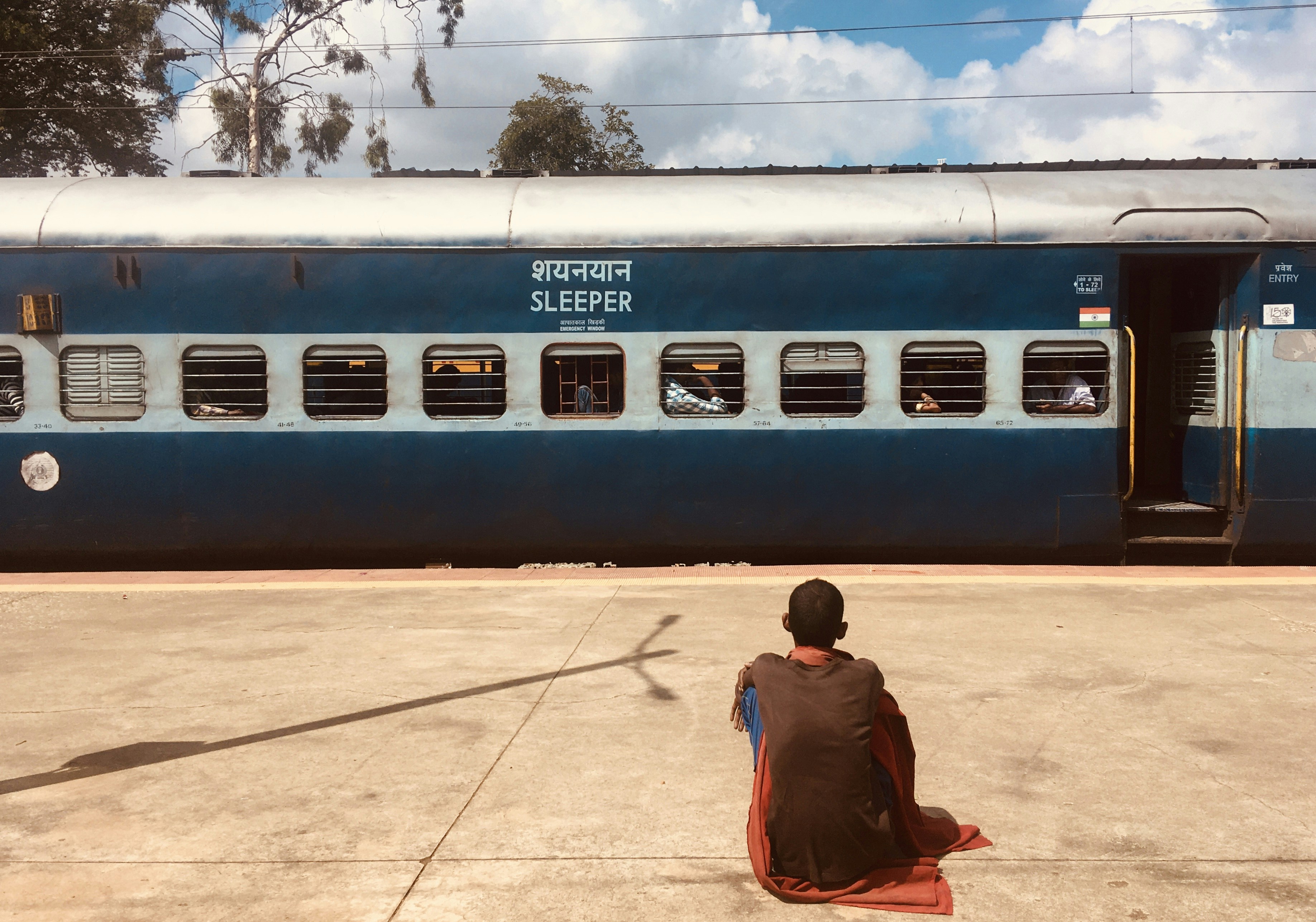 Mann in brauner Jacke sitzt tagsüber auf braunem Betonboden in der Nähe eines weißen und blauen Zuges