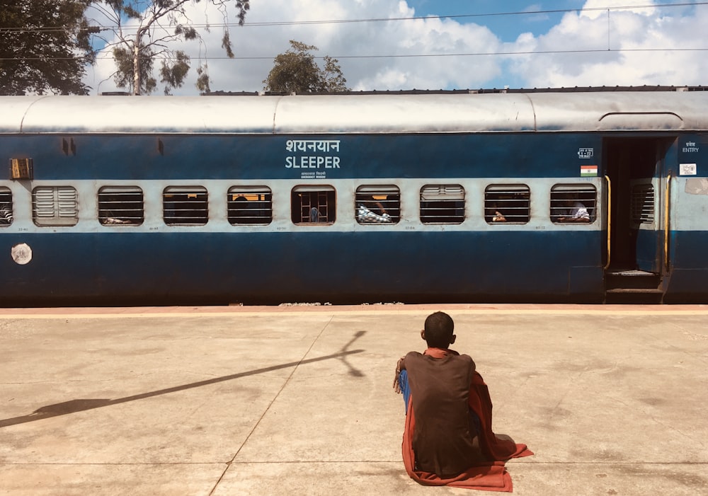 man in brown jacket sitting on brown concrete floor near white and blue train during daytime