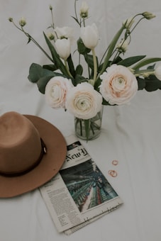 A bouquet of pale pink and white flowers in a glass vase is accompanied by a brown wide-brimmed hat resting on a newspaper. The setting includes small copper rings placed beside the newspaper on a white surface.