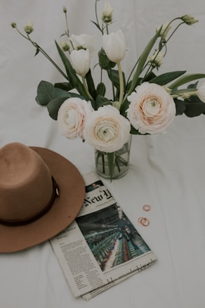 A bouquet of pale pink and white flowers in a glass vase is accompanied by a brown wide-brimmed hat resting on a newspaper. The setting includes small copper rings placed beside the newspaper on a white surface.