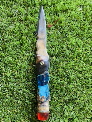 A handcrafted knife with a sharp, metallic blade and an intricately designed handle featuring various colors including blue and red. The knife is placed on a lush green grass background.