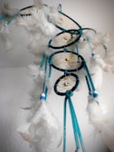 A dreamcatcher is suspended against a light-colored background, featuring a series of intricate circular patterns woven with threads. The structure is adorned with soft, white feathers and accented with small beads. Vibrant blue ribbons hang down, adding a striking contrast to the delicate feather arrangement.