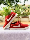 A pair of red sneakers with white soles and a white swoosh logo placed on a white towel. The background features a row of green trees and a residential building with a fence.