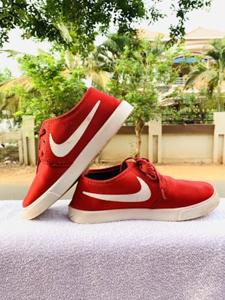A pair of red sneakers with white soles and a white swoosh logo placed on a white towel. The background features a row of green trees and a residential building with a fence.