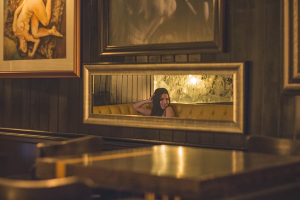 A warmly lit room with a reflective wall mirror captures the image of a person sitting on a yellow sofa. The background features dark wooden paneling and framed artwork, contributing to the intimate and cozy atmosphere.