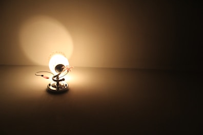 A decorative lamp with a floral design emits a soft, warm light. The lamp is positioned on a smooth surface, casting light and subtle shadows in a dimly lit room. The metal base and stem add a modern touch to the lamp's intricate design.