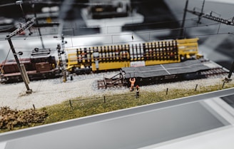A detailed model train setup on a display platform. The scene includes a model train track with a yellow train car and a dark brown engine. Tiny model figures dressed in bright orange workwear are positioned around the tracks, suggesting a construction or maintenance theme. Overhead wires and pylons are parts of the detail, adding realism to the miniature landscape.