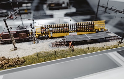A detailed model train setup on a display platform. The scene includes a model train track with a yellow train car and a dark brown engine. Tiny model figures dressed in bright orange workwear are positioned around the tracks, suggesting a construction or maintenance theme. Overhead wires and pylons are parts of the detail, adding realism to the miniature landscape.