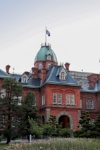 A historic red-brick building with a green domed roof stands prominently. The architecture is detailed, featuring arched windows and intricate brickwork. A flag flies atop the dome, and the building is surrounded by lush greenery, including several well-maintained trees and shrubs.
