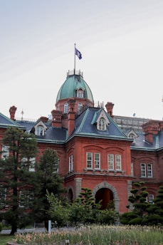 A historic red-brick building with a green domed roof stands prominently. The architecture is detailed, featuring arched windows and intricate brickwork. A flag flies atop the dome, and the building is surrounded by lush greenery, including several well-maintained trees and shrubs.