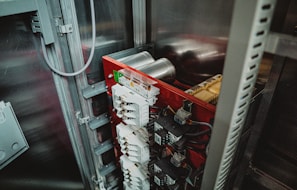 An electrical panel with several circuit breakers and transformers installed inside an industrial cabinet. The panel has a red backing and is surrounded by metal framing. Various wires and components are visible, indicating a complex setup.