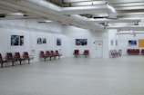 A spacious interior with red seating arrangements against white walls. Several framed pictures are displayed along the walls, and exposed ductwork is visible on the ceiling. The floor is made of light-colored material, enhancing the open and minimalist design.