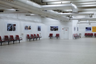 A spacious interior with red seating arrangements against white walls. Several framed pictures are displayed along the walls, and exposed ductwork is visible on the ceiling. The floor is made of light-colored material, enhancing the open and minimalist design.