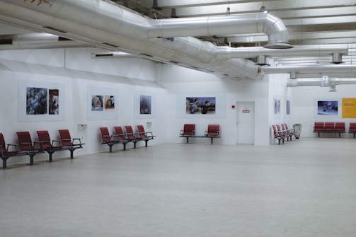 A spacious interior with red seating arrangements against white walls. Several framed pictures are displayed along the walls, and exposed ductwork is visible on the ceiling. The floor is made of light-colored material, enhancing the open and minimalist design.