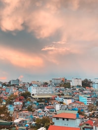 A vibrant urban landscape features a mix of colorful buildings with red, blue, and green rooftops. The architecture is eclectic, combining both modern and traditional styles. In the background, larger commercial buildings rise against a dramatic sky filled with soft orange and pink clouds, suggesting a sunset or sunrise.