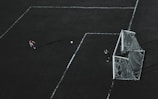 Aerial view of a soccer field with a player about to kick the ball towards the goal. A goalkeeper stands near the goal post ready to defend. The field markings are visible and the image has a dramatic lighting with long shadows.