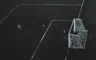 Aerial view of a soccer field with a player about to kick the ball towards the goal. A goalkeeper stands near the goal post ready to defend. The field markings are visible and the image has a dramatic lighting with long shadows.