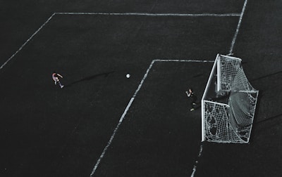 Aerial view of a soccer field with a player about to kick the ball towards the goal. A goalkeeper stands near the goal post ready to defend. The field markings are visible and the image has a dramatic lighting with long shadows.