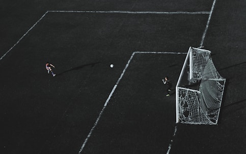 Aerial view of a soccer field with a player about to kick the ball towards the goal. A goalkeeper stands near the goal post ready to defend. The field markings are visible and the image has a dramatic lighting with long shadows.
