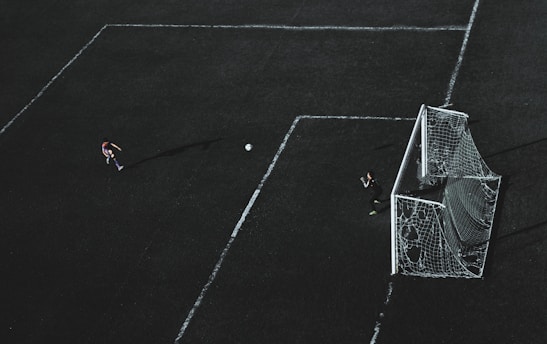Aerial view of a soccer field with a player about to kick the ball towards the goal. A goalkeeper stands near the goal post ready to defend. The field markings are visible and the image has a dramatic lighting with long shadows.