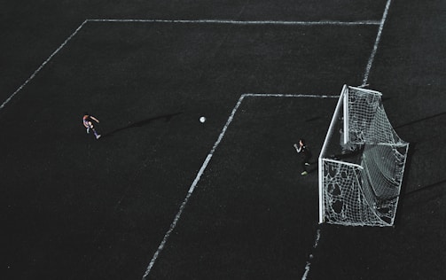 Aerial view of a soccer field with a player about to kick the ball towards the goal. A goalkeeper stands near the goal post ready to defend. The field markings are visible and the image has a dramatic lighting with long shadows.