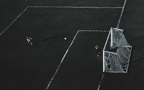 Aerial view of a soccer field with a player about to kick the ball towards the goal. A goalkeeper stands near the goal post ready to defend. The field markings are visible and the image has a dramatic lighting with long shadows.