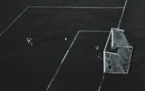 Aerial view of a soccer field with a player about to kick the ball towards the goal. A goalkeeper stands near the goal post ready to defend. The field markings are visible and the image has a dramatic lighting with long shadows.