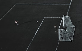 Aerial view of a soccer field with a player about to kick the ball towards the goal. A goalkeeper stands near the goal post ready to defend. The field markings are visible and the image has a dramatic lighting with long shadows.