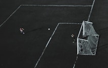 Aerial view of a soccer field with a player about to kick the ball towards the goal. A goalkeeper stands near the goal post ready to defend. The field markings are visible and the image has a dramatic lighting with long shadows.