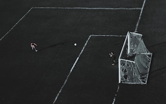 Aerial view of a soccer field with a player about to kick the ball towards the goal. A goalkeeper stands near the goal post ready to defend. The field markings are visible and the image has a dramatic lighting with long shadows.