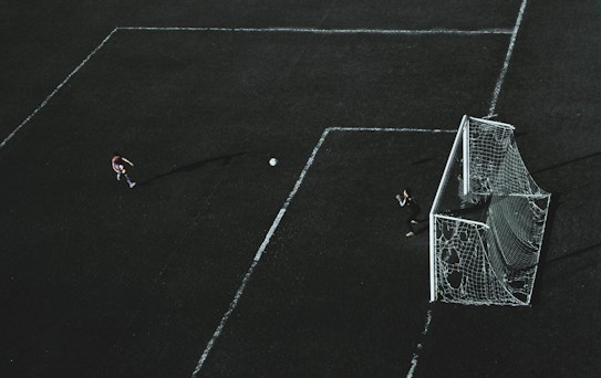 Aerial view of a soccer field with a player about to kick the ball towards the goal. A goalkeeper stands near the goal post ready to defend. The field markings are visible and the image has a dramatic lighting with long shadows.