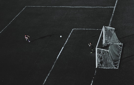 Aerial view of a soccer field with a player about to kick the ball towards the goal. A goalkeeper stands near the goal post ready to defend. The field markings are visible and the image has a dramatic lighting with long shadows.