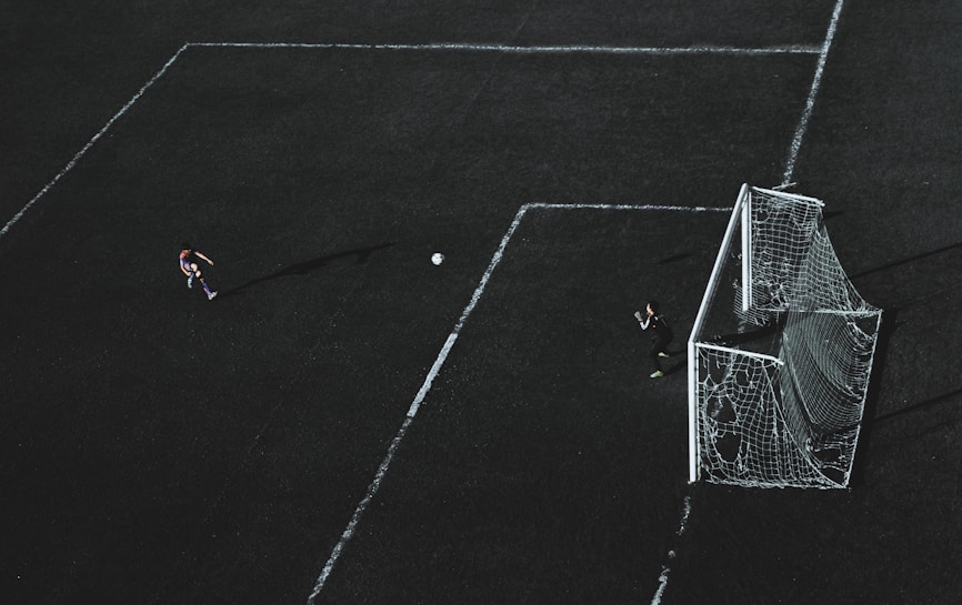 Aerial view of a soccer field with a player about to kick the ball towards the goal. A goalkeeper stands near the goal post ready to defend. The field markings are visible and the image has a dramatic lighting with long shadows.