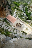 A small chapel with a red roof set in a rocky, mountainous landscape. Stone steps lead down to the chapel, surrounded by rich greenery and rugged terrain. A winding path continues into the distance, offering a sense of seclusion and tranquility.
